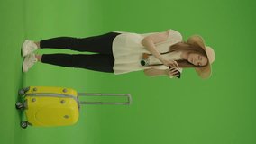 Vertical Video. Green Screen. Chroma Key. Woman Standing With a Hat and Luggage is Typing Something in Smartphone, Then raising Head and Starts Looking Away, and Continues Smiling. Satisfied Woman