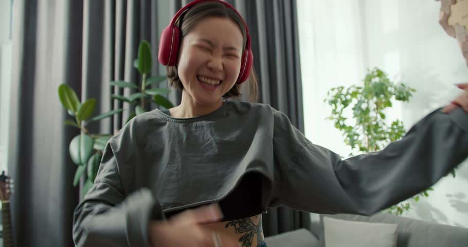 Asian woman playing air guitar and wearing headphones to listen to music, having fun at home Royalty-Free Stock Footage #1102964133