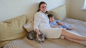 Baby with mother and cat watching TV sitting on sofa in home living room. Child, pet and mom sitting together on the couch