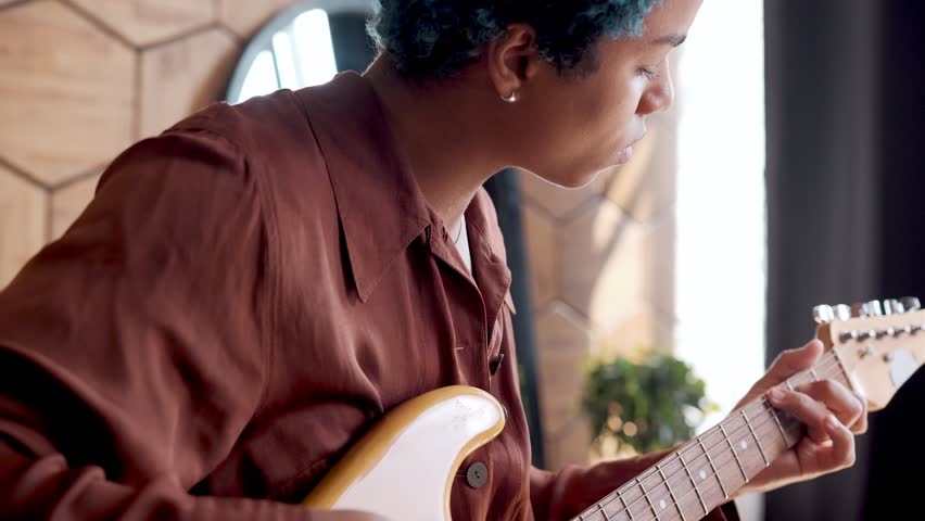 Young creative melancholic African American woman plays guitar and sings song recording audio on condenser microphone for posting track on Internet or on streaming service sits in home interior
