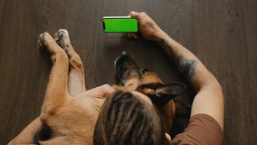 Owner resting with German Shepherd inside, stroking and hugging. Chroma Key Top view. Young man lying on wooden floor with dog uses smartphone to watch movie or video. Green screen, horizontal mockup.