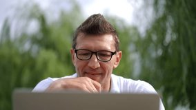 Front view close-up man in eyeglasses chatting online using web chat on laptop outdoors. Headshot portrait of intelligent confident Caucasian freelancer talking in slow motion