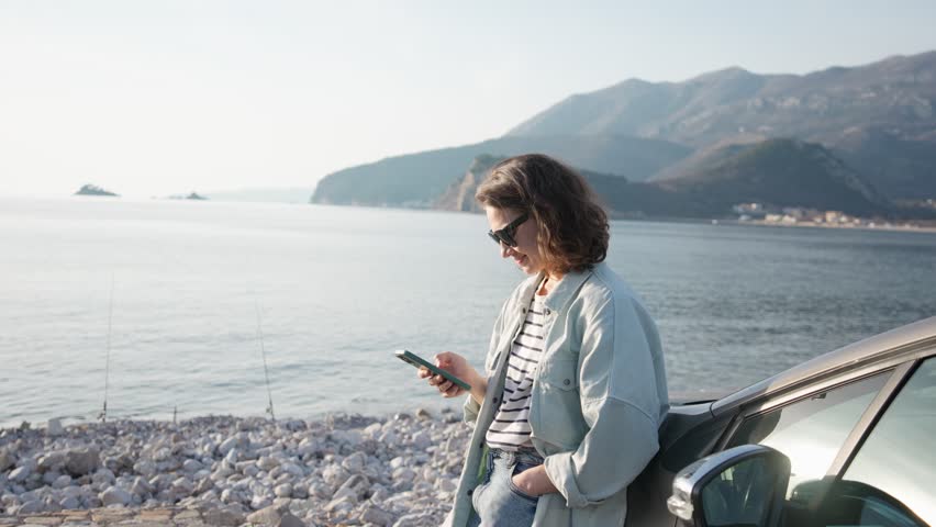 A young Caucasian woman using a phone leaning against her car parked with a beautiful view of the sea