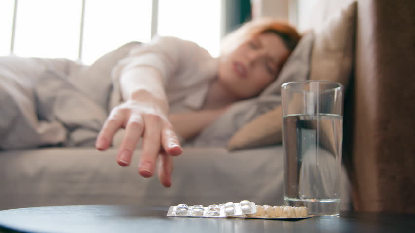 Glass of water and cure drugs on table in bedroom blurry sick woman suffering from headache in bed morning lady painful head migraine fever hangover pain flue coronavirus girl take pills painkiller | Shutterstock HD Video #1102976897