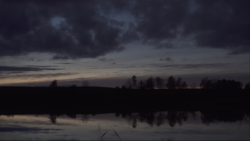 Nightfall time panorama at the lake with a dark cloudy sky reflecting on a water calm surface. Evening dusk romance with scenic clouds in heaven. Beautiful dark moods of the twilight hour at sundown.