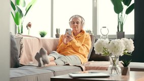 A beautiful middle-aged woman in comfortable clothes and wireless headphones is sitting on the sofa, holding the phone and listening to music, and moving to the music and being happy.