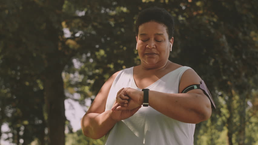 Medium slowmo of mid aged curvy African American woman setting up timer on her smartwatch while jogging outdoors in park at summertime Royalty-Free Stock Footage #1102991683