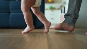 Father helps his son take his first steps on parquet floor. first awkward steps. Tiny legs gain confidence every moment. Joy of a father when he encourages his child to take another step. Happy family