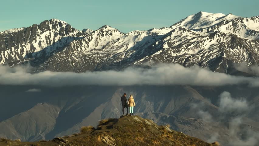 Couple standing on high peak with view of snow capped alpine mountains, aerial