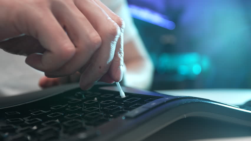 Extreme close-up of unrecognizable man cleaning dirty black keyboard disassembled from buttons using cotton bud sitting at table. Concept of general cleanup on workplace. Shooting in slow motion.