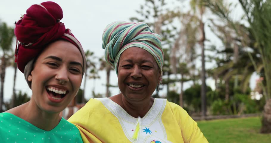 Happy african mother and daughter wearing traditional clothes sitting on meadow and smiling on camera with city park in background - Mom day and family concept
