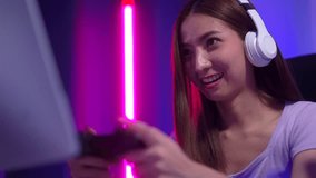 
Excite Young woman streamer wearing headphones playing game online and broadcast streaming studio in dark room neon light. Beautiful powerful female esports gamer play Cyber Gaming live at home.
