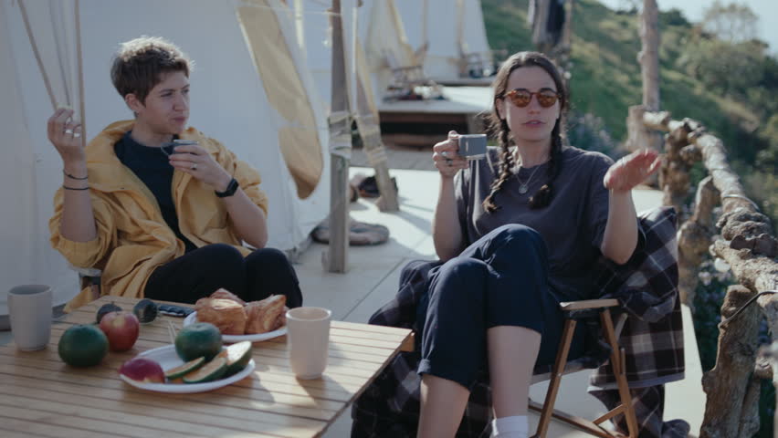 Two female friends sitting at glamping, having tea and meal, discussing the view of nature around. Weekend getaway, healthy recreational activity concepts