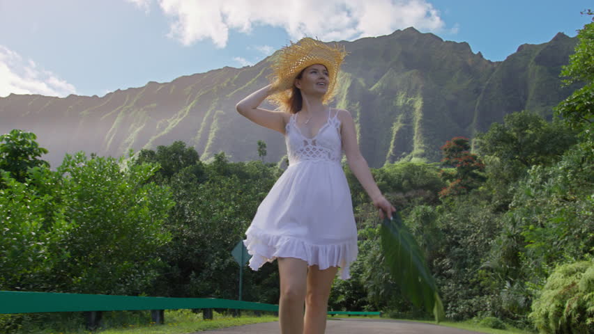 Attractive young woman 30s in white boho style mini dress walking by Hawaiian landscape, turns around with flying hair and feeling amazed by nature beauty. Happy relaxed lady enjoying Hawaii trip USA