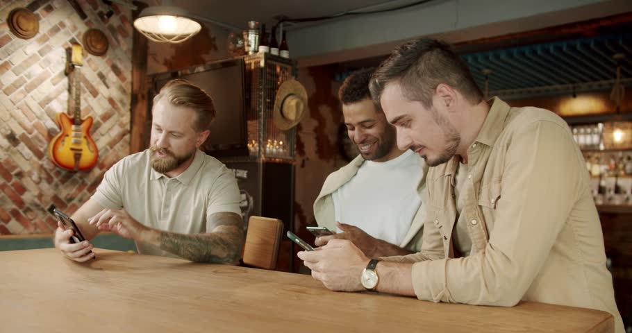 Looking at smartphones unconscious about the surroundings. Multiethnic group of people using their smartphones actively. Three men using their smartphones while sitting in pub after work. Royalty-Free Stock Footage #1103023679