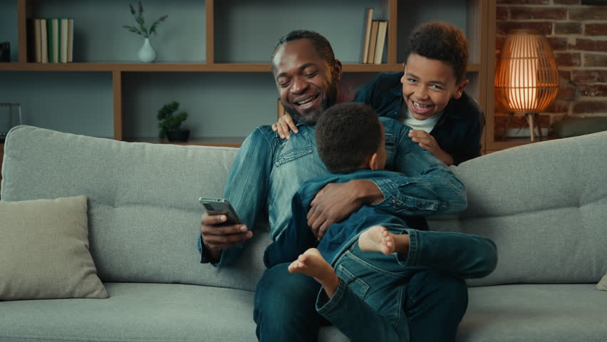 African American man father using mobile phone browsing smartphone at home couch sofa two little children kids sons boys abruptly suddenly appear embrace hugging daddy happy family bonding fatherhood Royalty-Free Stock Footage #1103025151