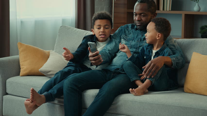 African American happy family using funny mobile phone app together at home having fun with smartphone service. Father with two sons little boys children on sofa browsing cellphone trying photo mask