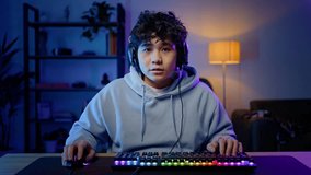 Young Asian male professional gamer wearing headphones, playing computer game, losing competition in hard match, looking at computer monitor, using computer mouse and keyboard. Cybersport, gaming club