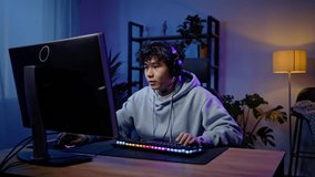 Slow motion of a young Asian man, professional gamer wearing headphones, sitting at computer monitor and nervously playing virtual online video game with winner expression at gaming room