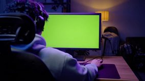 Selective focus on a computer monitor with green chroma key screen with free ad space for insert videos, at wooden desk on the background of a young man in audio headset, playing virtual online games