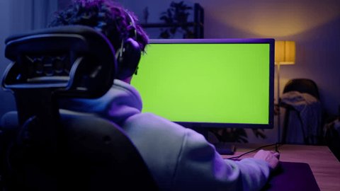Steadicam shot of a young man, computer gamer wearing headphones playing video games late at night, sitting at laptop with green chroma key screen with free space for inserting your advertising video วิดีโอสต็อก
