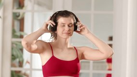 Sporty girl in tank top listening music in headphones and dancing at home. Woman enjoys music