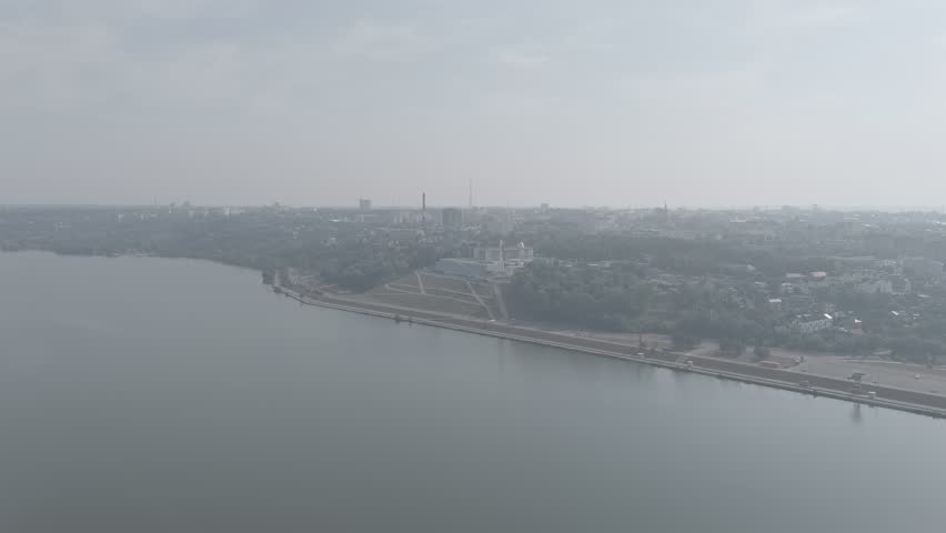Kaluga, Russia. The Tsiolkovsky State Museum of the History of Cosmonautics and its iconic Vostok rocket monument dominate this aerial view, set against the expansive Yachenskaya embankment and the calm waters of the Yachenskoye Reservoir. The museum building presents a striking modernist silhouette with its angular, white facade and dark glass panels, while the towering 38-meter Vostok rocket stands vertically on a raised granite pedestal, its metallic booster stages and distinctive conical shape clearly defined. The surrounding landscape features a mix of dense green foliage, manicured park areas, and a wide promenade that curves along the water