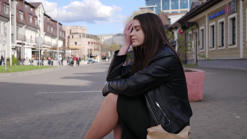 A sad beautiful girl sits on the curb on a city street