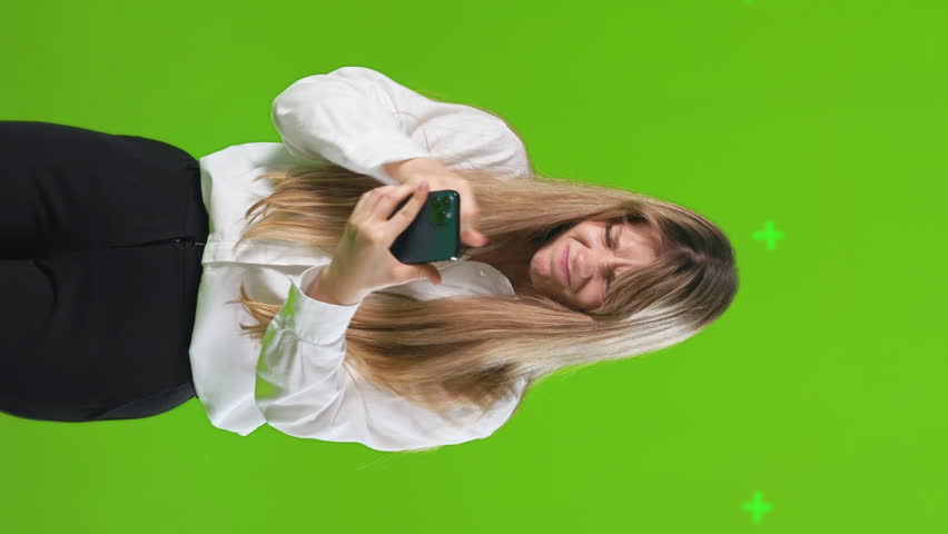 Upset young female feeling anger, while looking at phone screen in studio. Front view of irritated woman having problem with smartphone, isolated on green, horizontal. Concept of emotions, technology.