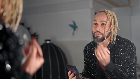 Gay man applying eyelashes looking mirror dressed black jacket with sequins . Brazilian black ethnicity homosexual guy with beard doing make up. Non-binary gender person. LGBT people gender identity Adlı Stok Video