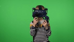 A boy in a virtual reality headset is playing a sword game on a green background. A boy in VR glasses and with joysticks in his hands is playing a virtual game. Chromakey.