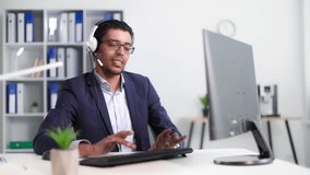 young successful black man in glasses and headphones with a microphone working at computer and typing on keyboards side in office