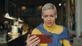 Caucasian mature lady sitting at modern cafe, drinking coffee and watching video on smartphone in earphones. Attractive female blonde wearing blue and yellow sweaters. People and technology