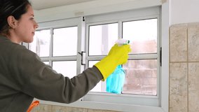Latin young woman cleaning windows with spray and rubber gloves. Close up video