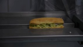 Close-up view of ciabatta sandwiches with chicken and vegetables moving on conveyer to packaging machine. Soft focus. Real time handheld video. Food production theme.