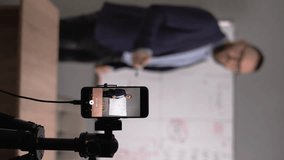 Tutor or English teacher stands near blackboard make speech on camera filming educational video for internet audience and his students. Vlogging, tutoring and education concept