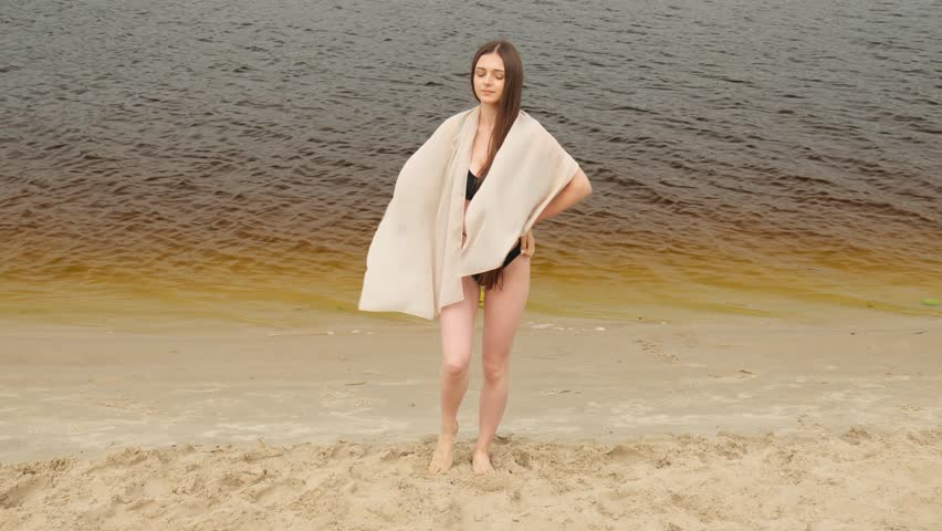A young girl with long hair in a dark swimsuit stands on the beach near the water with a gray towel. Slow motion beach. Relax summer