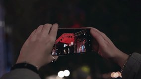 The girl shoots a night concert on stage on a mobile phone. Taking a close-up of her hand with the phone
