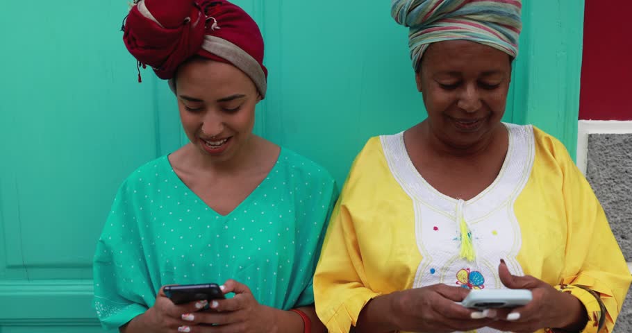 Happy african mother and her daughter wearing afro clothes using mobile phones in front of their house - Mom day, family and  culture concept