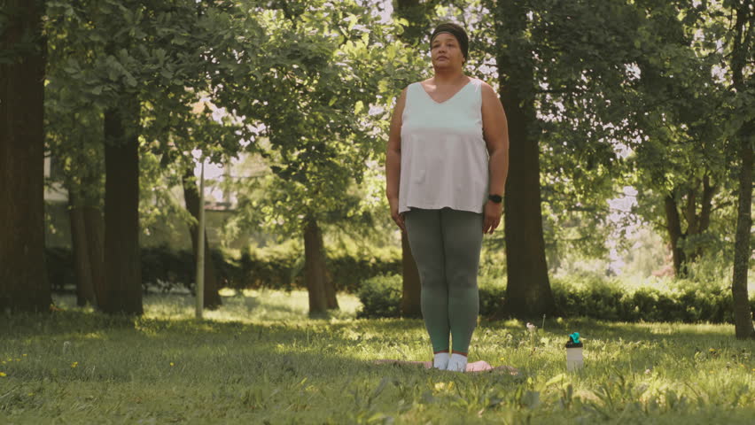 Full length slowmo of plus size African American mature woman meditating on green grass in summer park Royalty-Free Stock Footage #1103064359