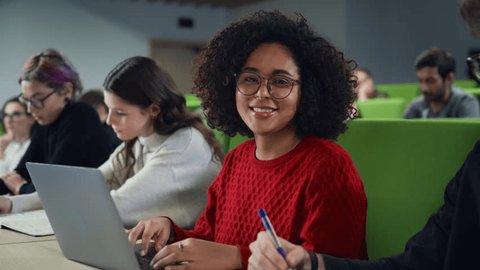 Portrait of an Empowered African Female Student Studying in University with Diverse Multiethnic Classmates. Young Happy Black Woman Looking at Camera and Smiling. Using Laptop Computer in Class : vidéo de stock