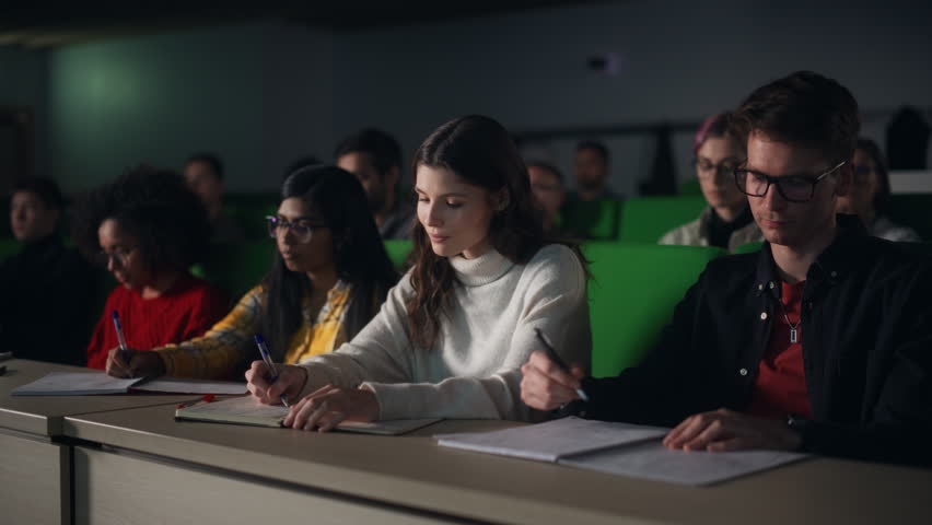Thoughtful Beautiful Young Female Studying in University, Listening to a Lecture in a Crowded Room with Diverse Multiethnic Classmates. Students Listening to Lecture and Taking Notes in Notebook