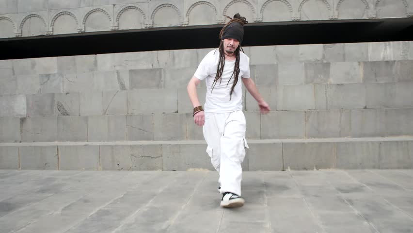 An attractive man in white clothes with African dreadlocks performs Brazilian capoeira movements and smiles sweetly. Young handsome man with beads around his neck and bracelets capoeira dance