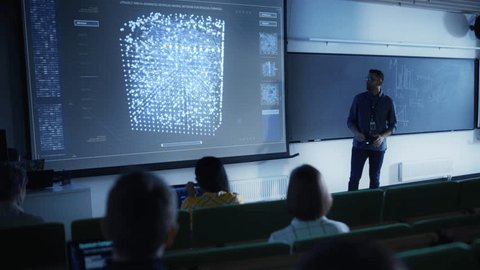 Young University Professor Explaining the Importance of Artificial Intelligence to a Group of Diverse Multiethnic Students in a Dark Auditorium. Teacher Showing Neural Network on Two Big Screens - Βίντεο στοκ