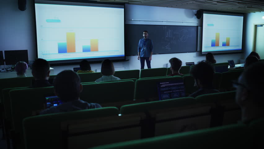Young Male Teacher Giving a Business Strategy Lecture to Diverse Multiethnic Group of Female and Male Students in Dark College Room. Projecting Slideshow with Comparison Charts and Commercial Data Royalty-Free Stock Footage #1103067037