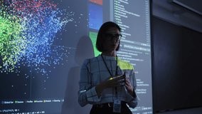 Young Female Teacher Giving a Data Science Presentation in a Dark Auditorium with Projecting Slideshow with Artificial Intelligence Neural Network Architecture. Business Startup and Education Concept - Powered by Shutterstock - Get 15% off with code: PIKWIZARD15