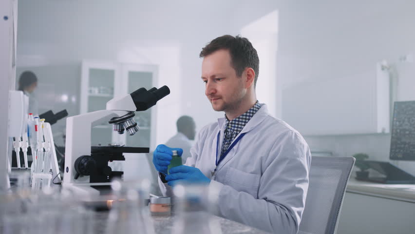 Shot of serious focused young Caucasian man wearing white lab coat sitting in front of modern microscope and holding lab jar with green substance in laboratory. Working day. Research concept Royalty-Free Stock Footage #1103070485