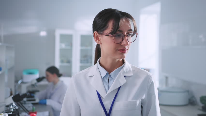 Close-up portrait of good-looking female biologist in glasses and white lab coat looking at camera in modern laboratory room. Young Caucasian woman posing in workplace. Profession, job concept Royalty-Free Stock Footage #1103070519