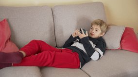 Child lying on the sofa watching video or photo content in social network, scrolling by smart phone, smiling boy enjoys mobile app at home. Online education, communicating distantly, playing games