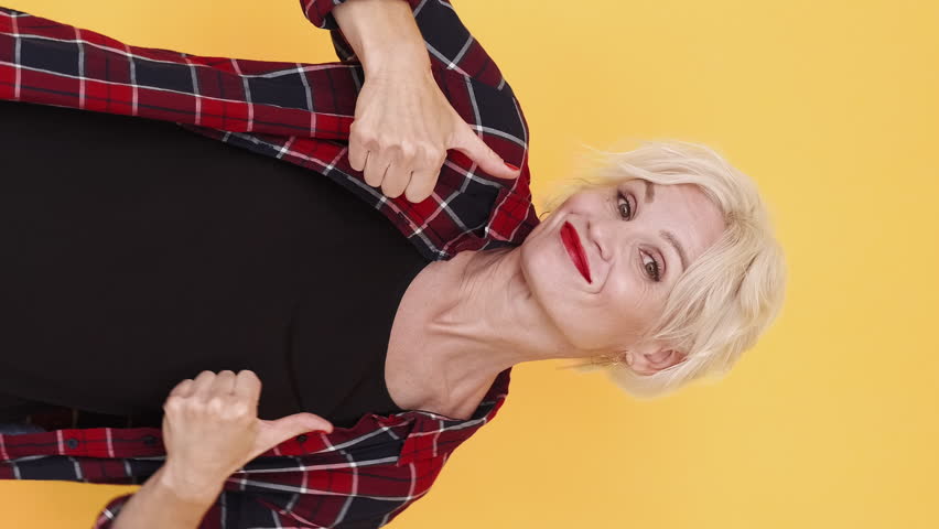 Vertical video. Many likes. Gesturing woman. Approving joy. Happy middle-aged lady showing thumbs up dancing feeling good on yellow.